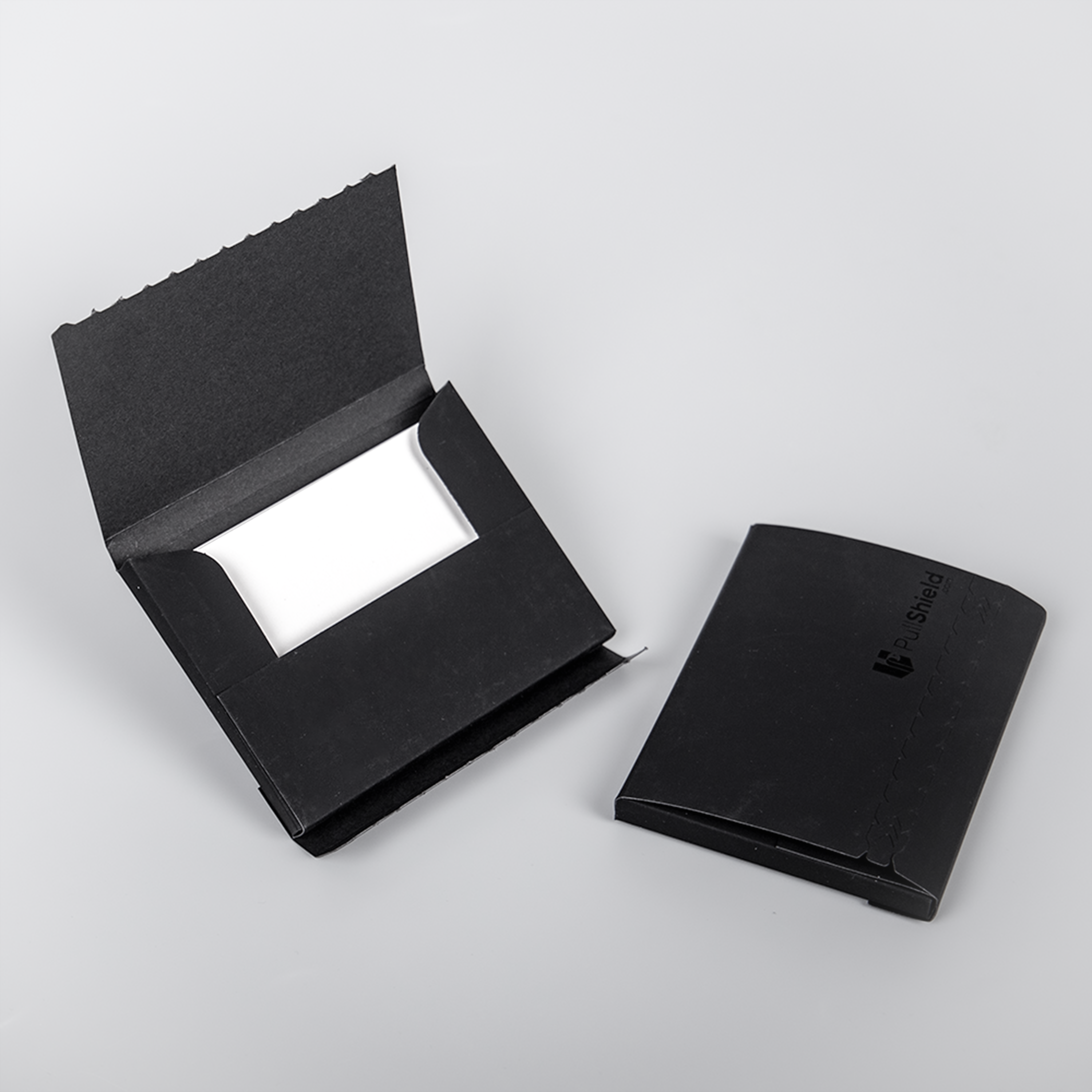 “Two black PullShield protective TCG card sleeves on a light gray background. One sleeve is open, showing the inner white cardboard insert, while the other sleeve is sealed and closed with the PullShield logo visible.
